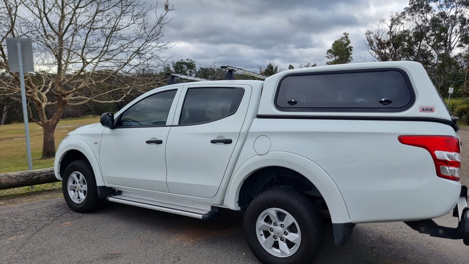 A white Mitsubishi Triton dual-cab ute with a canopy and roof racks, parked beside a grassy reserve under a grey sky